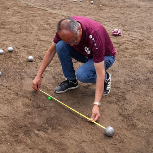 Ein Mann misst mit einem Ma&szlig;band den Abstand zwischen einer gr&uuml;nen Kugel und mehreren silbernen Kugeln auf einem Sandplatz.