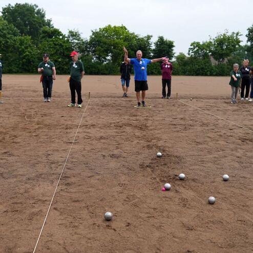 Gruppen von Menschen auf einem Sandplatz, w&auml;hrend sie Boule-B&auml;lle werfen und die Spielpositionen beobachten.