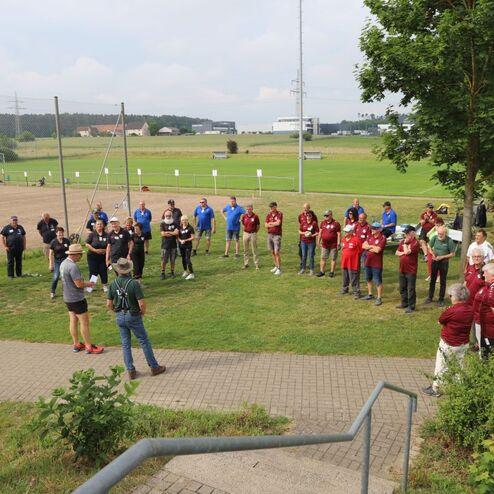 Gruppentreffen von Sportlern im Freien, einige h&ouml;ren einem Redner auf einer Wiese zu, andere stehen in der N&auml;he.