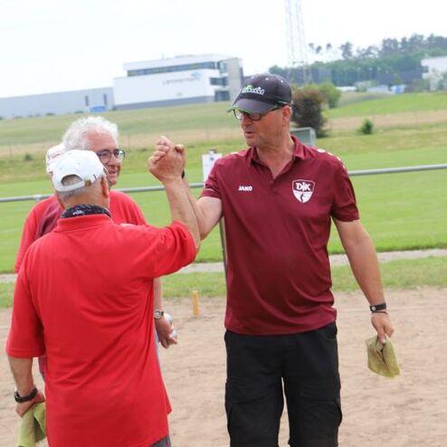 Drei M&auml;nner in rot und braun stehen auf einem Sportplatz und sch&uuml;tteln sich l&auml;chelnd die H&auml;nde.