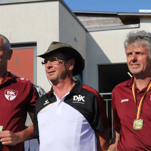 Drei M&auml;nner in Sportkleidung stehen zusammen, einer tr&auml;gt einen Hut, alle halten Medaillen und eine Urkunde.