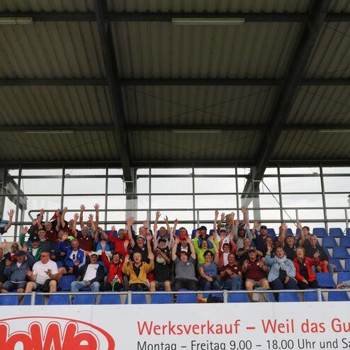 Gruppe von Menschen auf einer Trib&uuml;ne, viele winken fr&ouml;hlich und tragen verschiedene farbige Kleidung.