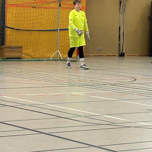 Junger Torwart in leuchtend gelber Kleidung steht vor einem Handballtor in einer Sporthalle.