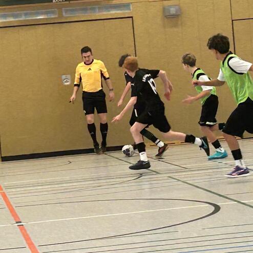 F&uuml;nf Jugendliche spielen Fu&szlig;ball in einer Sporthalle, ein Schiedsrichter begleitet das Spiel.
