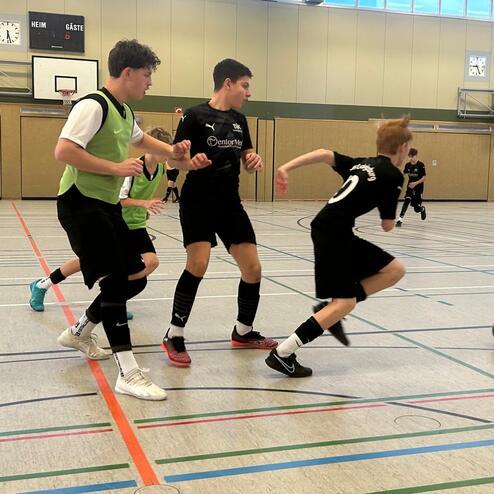 Zwei Spieler im Schwarz k&auml;mpfen um den Ball, w&auml;hrend ein dritter Spieler in Gr&uuml;n von der Seite kommt. Halle mit Sportboden.