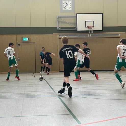 Fussballspiel in einer Halle mit mehreren Spielern in gr&uuml;nen und schwarzen Trikots, ein Ball rollt &uuml;ber den Boden.