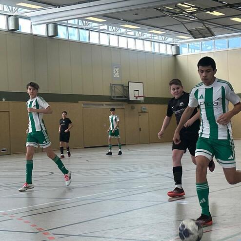 Drei Jugendliche spielen Fu&szlig;ball in einer Sporthalle; einer dribbelt den Ball, w&auml;hrend die anderen in Bewegung sind.