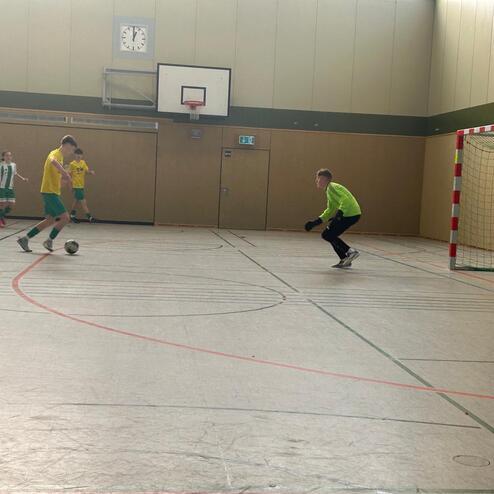 Zwei Fu&szlig;ballspieler dribbeln den Ball auf einem Hallenfu&szlig;ballfeld, w&auml;hrend der Torwart sich positioniert.