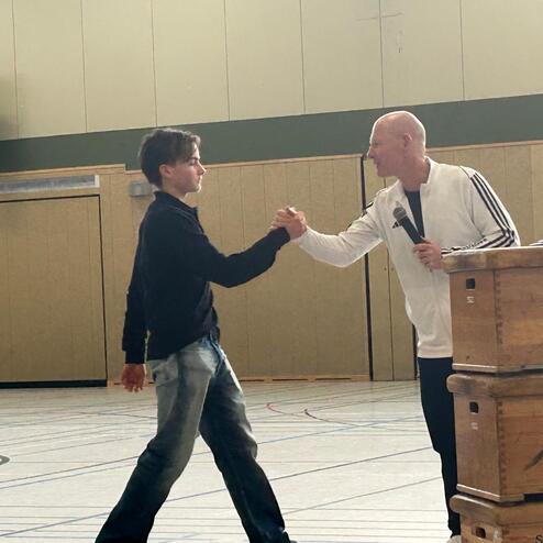 Zwei M&auml;nner geben sich in einer Sporthalle die Hand, w&auml;hrend einer ein Mikrofon h&auml;lt. Gymnastikger&auml;te sind im Hintergrund sichtbar.