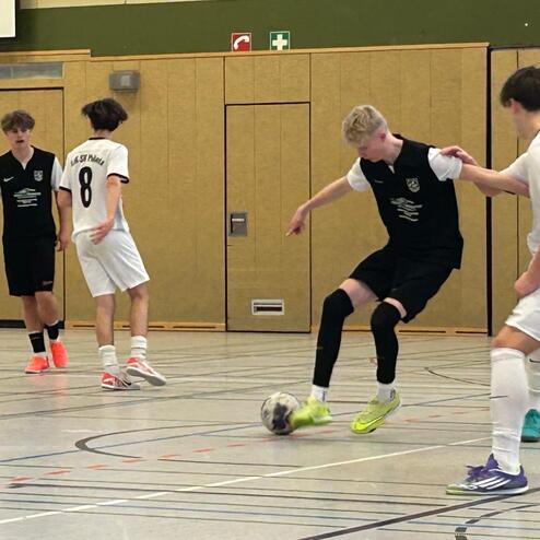 Zwei Fu&szlig;ballspieler dribbeln den Ball in einer Sporthalle, w&auml;hrend zwei weitere Spieler in der N&auml;he stehen.