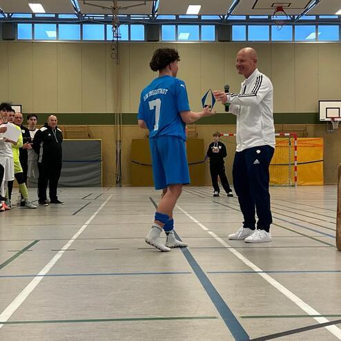 Ein Junge im blauen Trikot &uuml;berreicht einen Preis an einen Erwachsenen in Sportkleidung in einer Sporthalle.