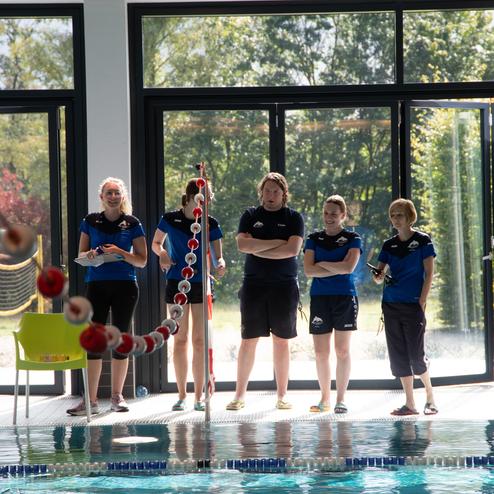 F&uuml;nf Personen in blauen Shirts stehen am Beckenrand eines Schwimmbads mit gro&szlig;en Fenstern und Blick ins Freie.