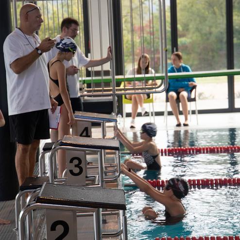Trainer gibt Anweisungen an Schwimmerin, w&auml;hrend andere Sportler am Beckenrand auf den Wettkampf warten.