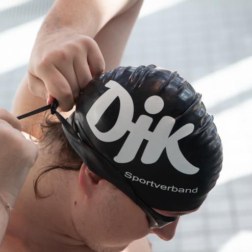Schwimmer bindet einen schwarzen Badekappenriemen mit dem Logo &bdquo;DJK Sportverband&ldquo; vor einem Fliesenboden.