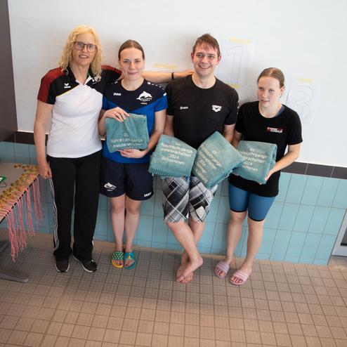 Gruppenfoto von vier Personen, die Schwimmpreise halten, vor einem Hallenbad mit Medaillen im Hintergrund.