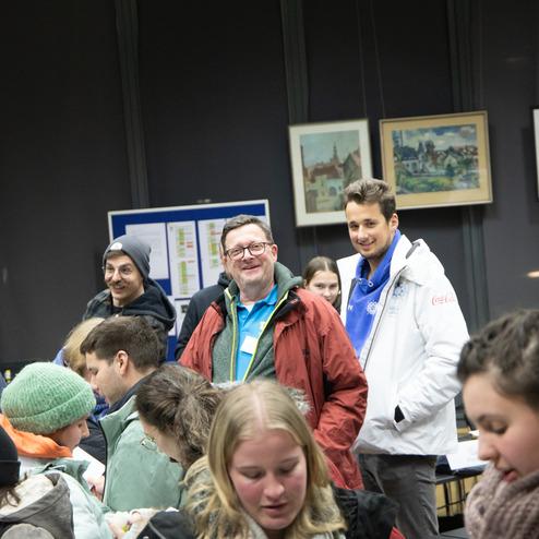 Gruppenszene mit mehreren Personen, die in einem Raum miteinander interagieren, umgeben von Bildern und Luftballons.