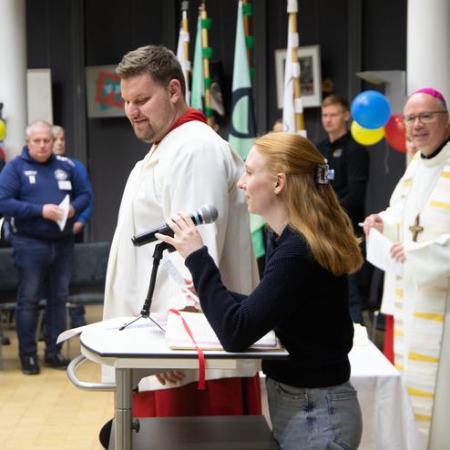 Eine Frau spricht mit Mikrofon, w&auml;hrend ein Mann in liturgischer Kleidung und ein Bischof aufmerksam zuh&ouml;ren.