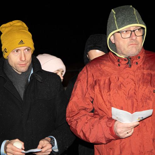 Zwei M&auml;nner mit Winterkleidung stehen im Freien, einer in gelbem M&uuml;tze und schwarzem Anzug, der andere in rotem Mantel.