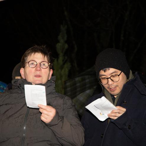 Zwei Personen stehen im Freien, halten Liederbl&auml;tter und singen in einer Gruppe von Menschen bei Nacht.