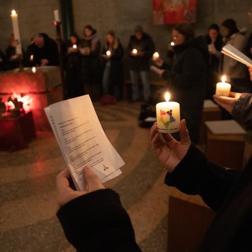 Menschen halten Kerzen und Gesangsb&uuml;cher in einem dunklen Raum w&auml;hrend einer Zeremonie mit stimmungsvollem Licht.