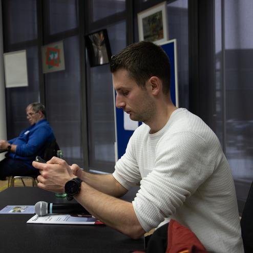 Ein Mann in einem wei&szlig;en Pullover tippt auf seinem Smartphone an einem Tisch, w&auml;hrend im Hintergrund eine andere Person sitzt.