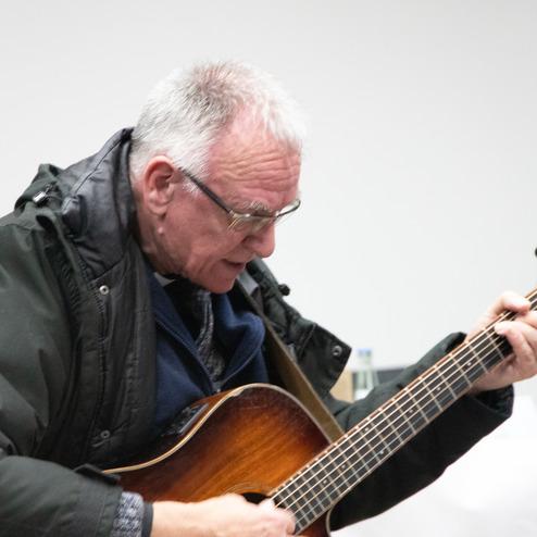 &Auml;lterer Mann mit Brille spielt eine Gitarre in einem Innenraum.