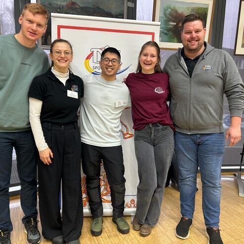 Gruppenbild von f&uuml;nf Personen, die vor einem Banner einer Sportorganisation stehen, l&auml;chelnd und in Freizeitkleidung.
