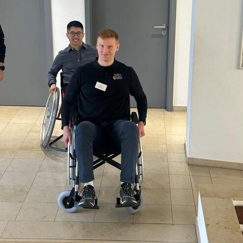 Zwei Personen in Rollst&uuml;hlen fahren eine Treppe hinunter; eine Person im Vordergrund, eine im Hintergrund.