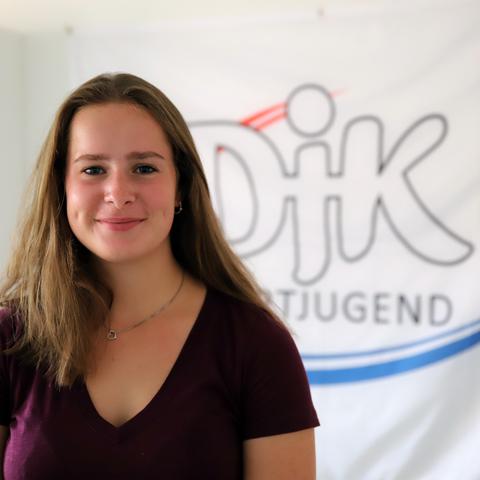 Portr&auml;t einer l&auml;chelnden jungen Frau vor einem Banner der DJK Jugend. Sie tr&auml;gt ein lila Oberteil und hat lange, braune Haare.