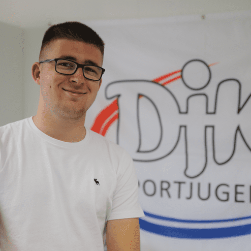 Junger Mann mit Brille in T-Shirt vor einem Banner der DJK Sportjugend.