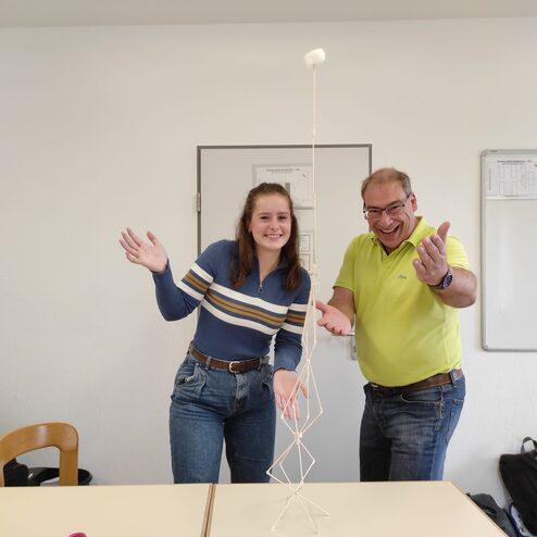 Zwei Personen l&auml;cheln und zeigen auf einen hohen Turm aus Spaghetti und einem Marshmallow auf einem Tisch.