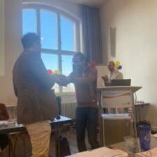 Zwei Personen sch&uuml;tteln sich die H&auml;nde w&auml;hrend einer Veranstaltung, im Hintergrund bunte Luftballons und ein Pr&auml;sentationstisch.
