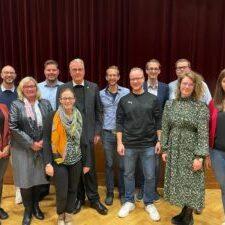 Gruppenfoto von 12 Personen vor rotem Vorhang, l&auml;chelnd, in verschiedenen Kleidungsstilen und Posen.