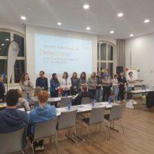 Gruppe von Personen steht an einem Tisch in einem hellen Raum, w&auml;hrend eine Pr&auml;sentation im Hintergrund gezeigt wird.