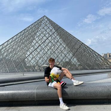 Junger Mann sitzt mit einem Pl&uuml;schtier vor der Glaspyramide des Louvre unter blauem Himmel.