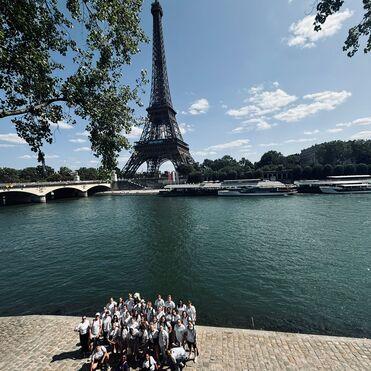 Gruppe von Menschen posiert am Ufer der Seine vor dem Eiffelturm an einem sonnigen Tag.