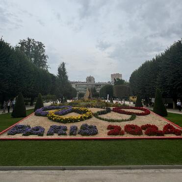 Blumenbeet mit dem Schriftzug &bdquo;Paris 2024&ldquo; und olympischen Ringen in einem Park, umgeben von B&auml;umen und Besuchern.
