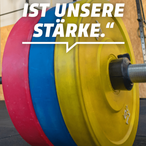 Nahaufnahme von Gewichten in den Farben Blau, Rot und Gelb auf einer Hantelstange, mit Schriftzug "Vielfalt ist unsere St&auml;rke."