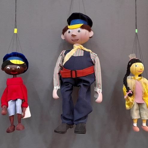 Drei h&auml;ngende Handpuppen: ein Junge in Uniform, ein j&uuml;ngerer Junge in Rot und ein M&auml;dchen mit buntem Outfit.