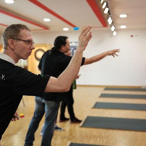 Drei M&auml;nner spielen Dart in einem Turnierraum, w&auml;hrend einer gerade seinen Pfeil auf die Zielscheibe wirft.