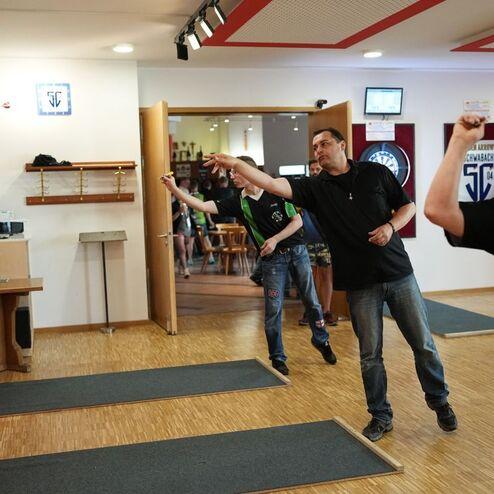 Drei M&auml;nner werfen Pfeile auf Dartboards in einem gut beleuchteten Spielraum mit Holzboden.