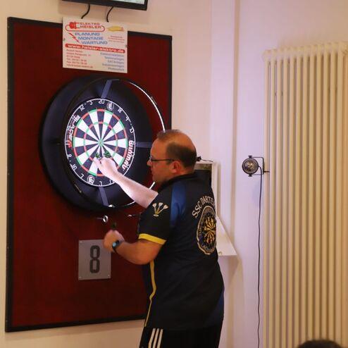 Mann mit Brille wirft einen Dartpfeil auf eine Dartscheibe an der Wand in einem Vereinsraum.