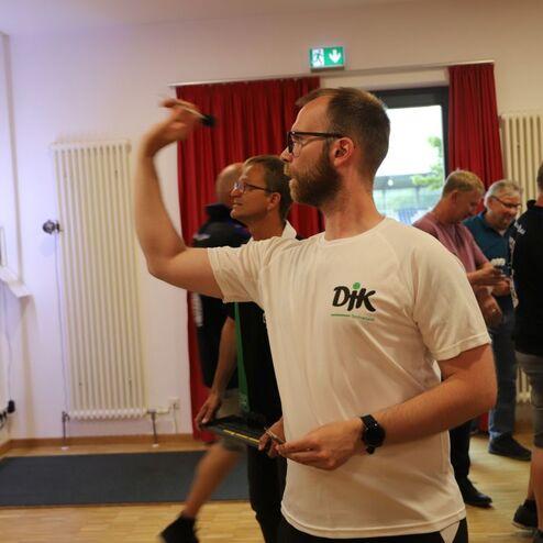 Mann mit Brille wirft Dartpfeil auf eine Dartscheibe in einem hellen Raum mit anderen Spielern im Hintergrund.