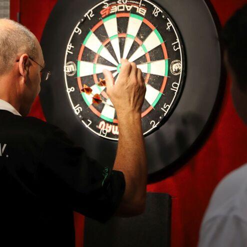 Mann wirft Pfeil auf eine Dartscheibe, hinter ihm ein zweiter Mann in einem wei&szlig;en Shirt.