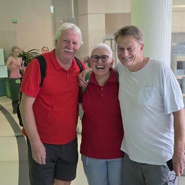 Drei Personen in roten Shirts stehen lächelnd in einem hellen Raum mit Tischen und Stühlen.