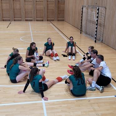 Mannschaftssitzung einer Handballmannschaft mit Trainer in der Halle, Spielerinnen sitzen im Kreis auf dem Boden.