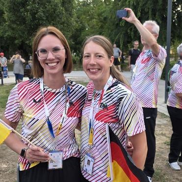Zwei Frauen in farbenfrohen T-Shirts halten eine deutsche Flagge und lächeln in einer feierlichen Umgebung.