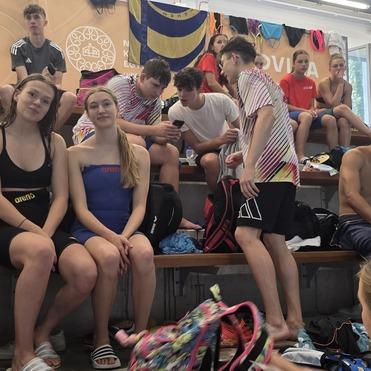Jugendliche schwimmen und sitzen in einem Schwimmhalle auf Tribünen, einige tragen Badebekleidung, andere sportliche Outfits.