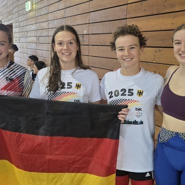 Vier Jugendliche halten deutsche Flaggen und posieren in einer sportlichen Umgebung mit Holzverkleidung im Hintergrund.