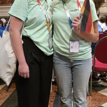 Zwei junge Frauen in grünen T-Shirts mit „VOLUNTEER“-Aufdruck halten eine kleine Deutschlandflagge in einem festlichen Raum.